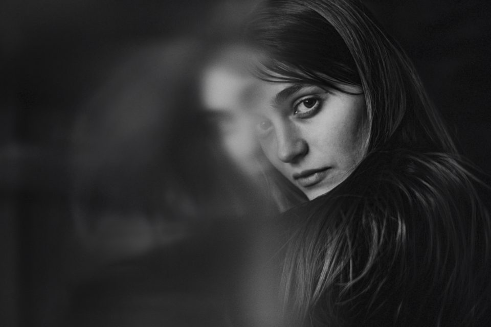 Black and white photo of a woman with long hair looking directly at the camera. She is partially obscured by a soft blur, creating a mysterious and contemplative atmosphere.