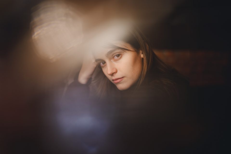 A woman with long hair gazes thoughtfully through a blurred, reflective surface with her head resting on her hand. The image has an ethereal, soft-focus effect.