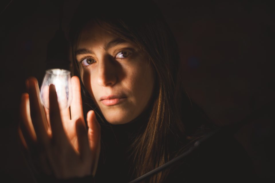 A person with long hair holds a lit bulb close to their face, illuminating their features in a dimly lit environment.