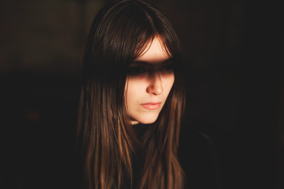 A person with long brown hair and a serious expression, partially illuminated with shadows casting across their face, looking slightly to the side.