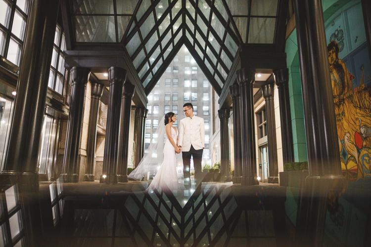 A couple in formal attire stand holding hands under a glass canopy, illuminated by city lights in the background. Captured with the precision of a Canon EOS R, this stunning moment showcases night photography's allure, perfect for timeless wedding portraits.