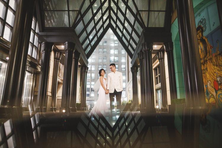A couple in formal attire stands holding hands under a modern glass-covered structure with city lights in the background, captured beautifully with a Canon EOS R and 24mm f/1.4 lens, perfect for wedding portraits.