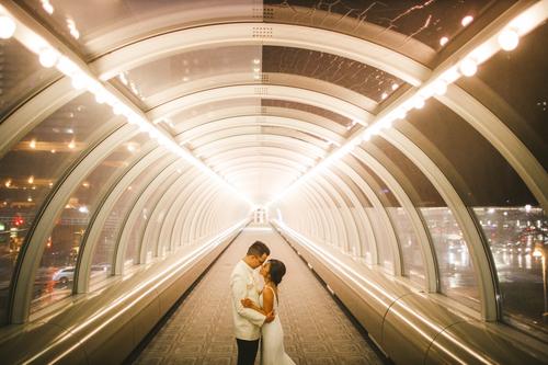 A couple stands close, embracing in the center of a brightly lit, futuristic-looking glass tunnel at night—a perfect setting for wedding portraits captured with the Canon EOS R and a 24mm f/1.4 lens.