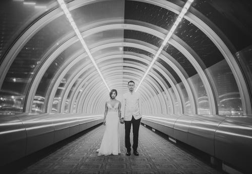 A couple stands holding hands in a well-lit, futuristic tunnel with curved transparent walls. Captured using a Canon EOS R with a 24mm f/1.4 lens, the woman is wearing a white dress, and the man is dressed in a white shirt and dark pants, making for perfect wedding portraits.