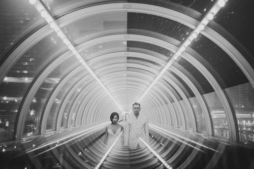 A man and a woman walk through a well-lit, transparent tunnel with lights reflected on the curved walls, perfect for capturing wedding portraits. The background showcases a vibrant cityscape with buildings and lights, ideal for night photography with the Canon EOS R.