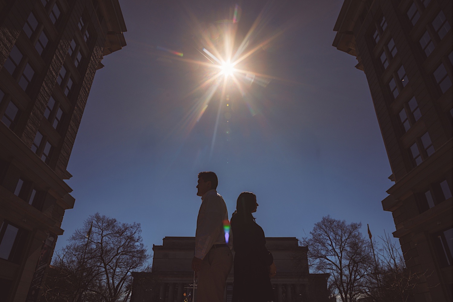 Two individuals stand back-to-back, silhouetted against the sun between two tall buildings, with bare trees and a distant structure in the background. It's a scene reminiscent of Joe Test's striking compositions.