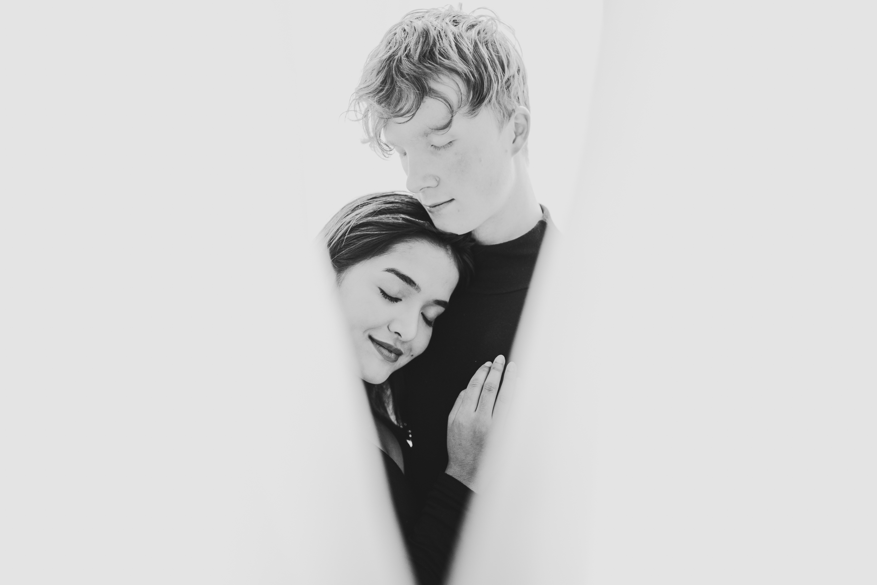 A couple embracing, woman leaning her head on man's chest, seen through a narrow, white-framed opening, in high contrast black and white. For FAQ regarding this image, please contact us.