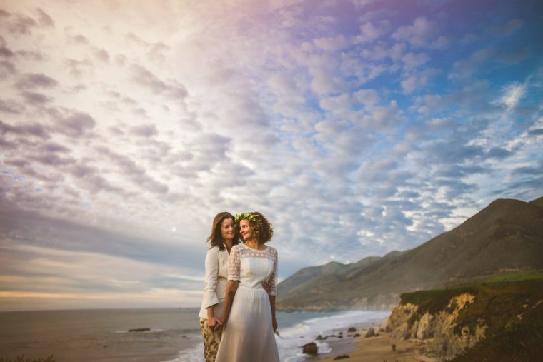 Two brides holding hands on a beach at sunset, with dramatic clouds overhead and mountains in the background.
