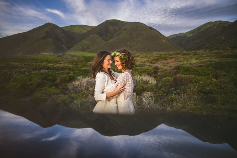 Two women embracing in a field with their reflection visible in a still pond, flower crowns in their hair, with hills in the background.