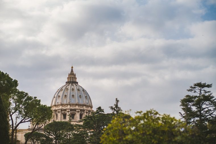 A distant view of St. Peter's Basilica dome peeking through lush foliage under a partly cloudy sky.