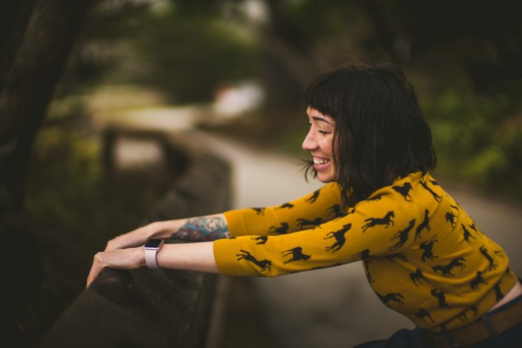 Woman in a yellow shirt with horse prints leaning on a wooden railing, smiling and looking to the side, surrounded by greenery.