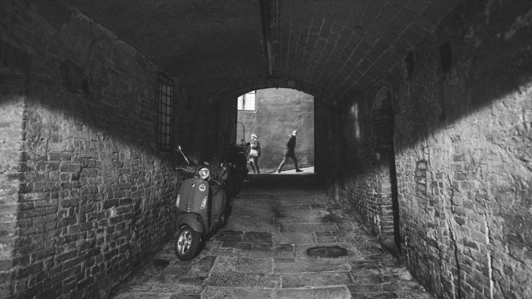 Black and white image of a brick tunnel with a scooter on the left side and two people standing at the far end.