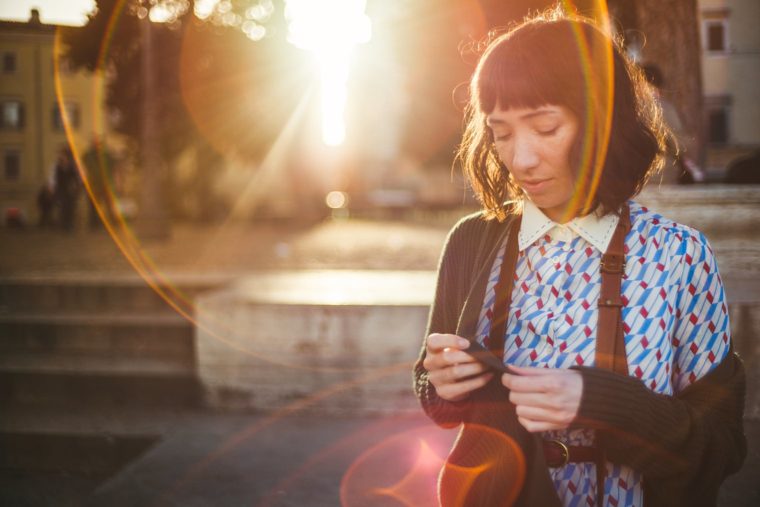 Woman with shoulder-length hair standing outdoors at sunset, looking down at her hands, with a lens flare effect in the background.