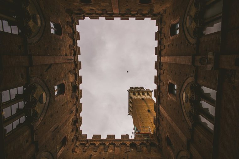 View from a courtyard looking up through a square opening, framed by building walls, with a tower visible against a cloudy sky and a bird flying.