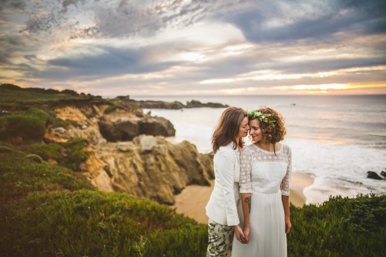 Two women in wedding attire affectionately touching foreheads on a coastal cliff at sunset, with clouds and ocean in the background.