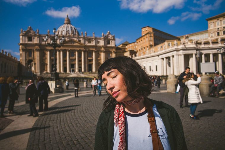 A young woman smiling with closed eyes in front of St. Peter's Basilica in Vatican City, with tourists in the background.