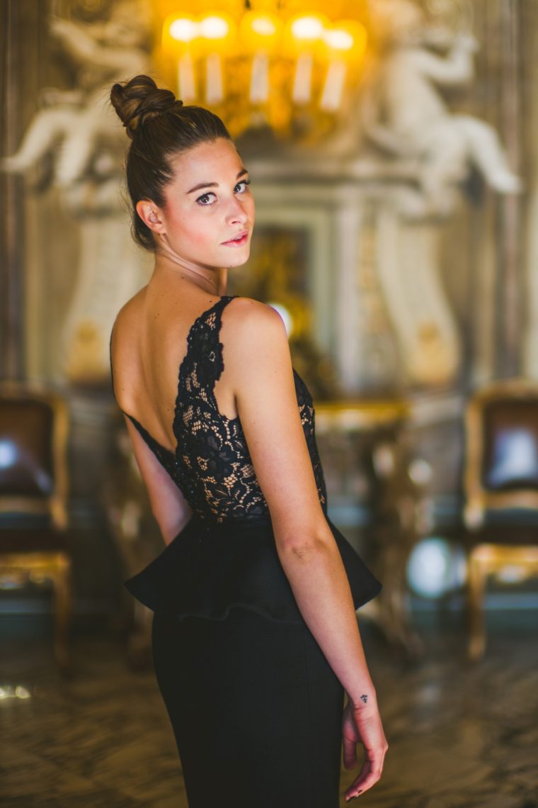 A woman in an elegant black lace dress looks over her shoulder in a richly decorated room with gilded details and chandeliers.
