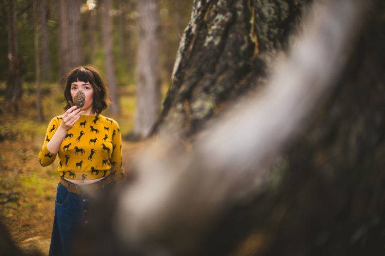 A young woman in a yellow shirt holding a pine cone to her face stands in a forest, partially obscured by a blurred foreground tree branch.