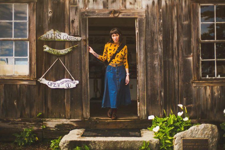 A woman in vintage clothing stands in the doorway of a rustic wooden cabin marked as "Halper's Cabin Museum.