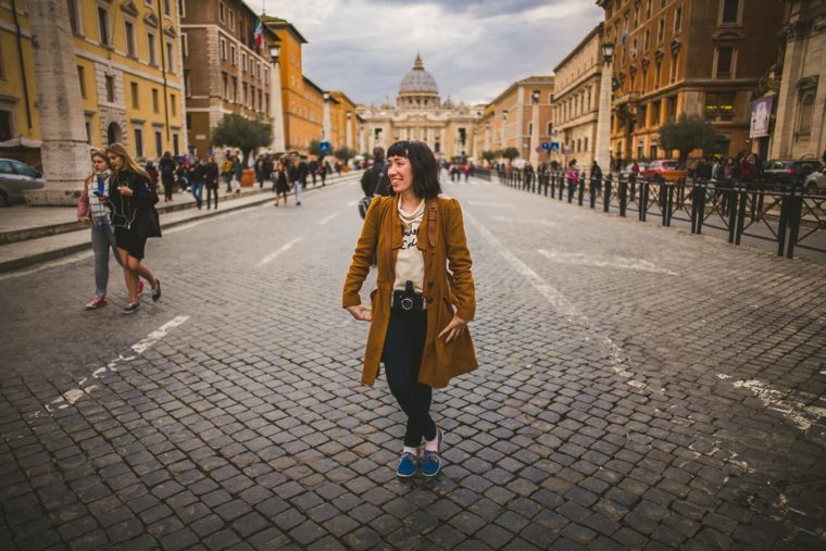 A woman in a tan coat and blue jeans smiles while walking across a cobblestone street, with a historic city scene and a dome in the background.