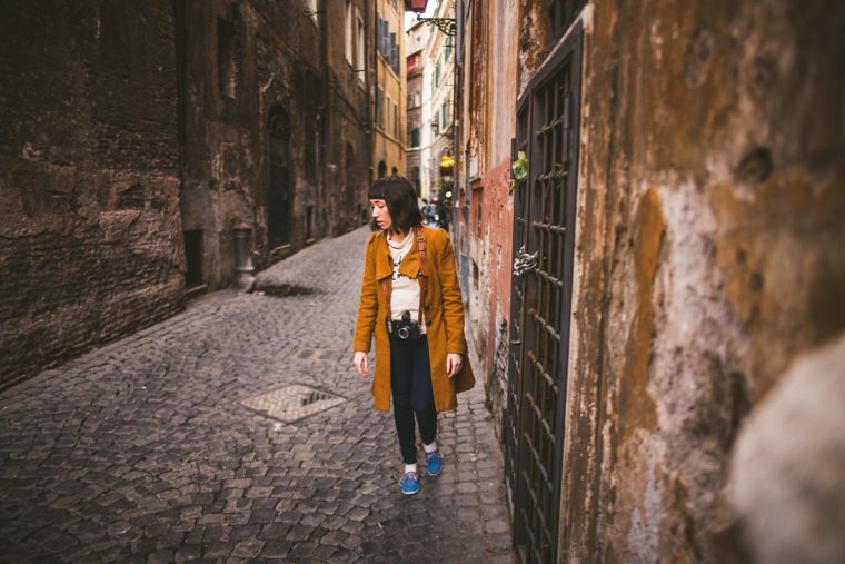 A person walks down a narrow, cobblestone street lined with old buildings. The person is wearing a long brown coat, holding a camera, and looking to the side.