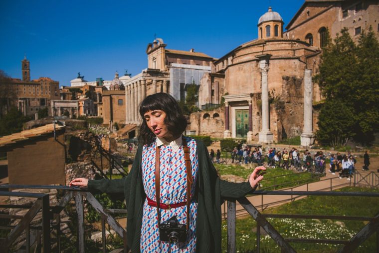 Woman with camera standing in front of ancient Roman buildings on a sunny day, with tourists in the background.