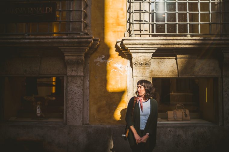 A woman stands in a sunlit European street, with a patterned shadow casting across an old building and display windows behind her.