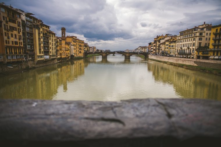 View of the Arno River with Ponte Vecchio in the distance, flanked by historic buildings under a cloudy sky, seen from behind a wooden ledge.