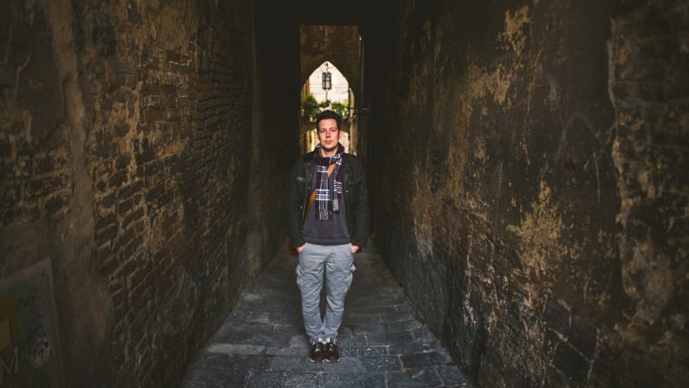 A person stands in a narrow, dimly lit alleyway with rugged brick walls and an opening at the end lit by daylight.