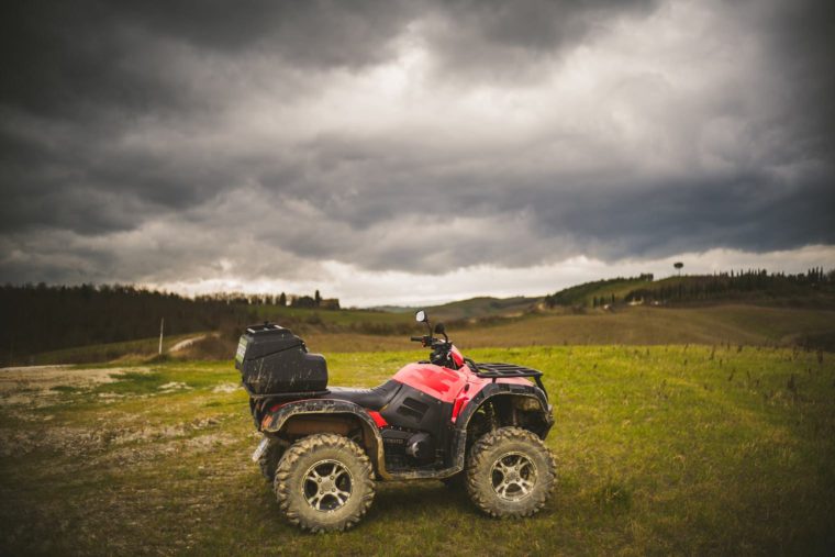 Red ATV parked on a grassy hill with a cloudy sky overhead and rolling countryside in the background.