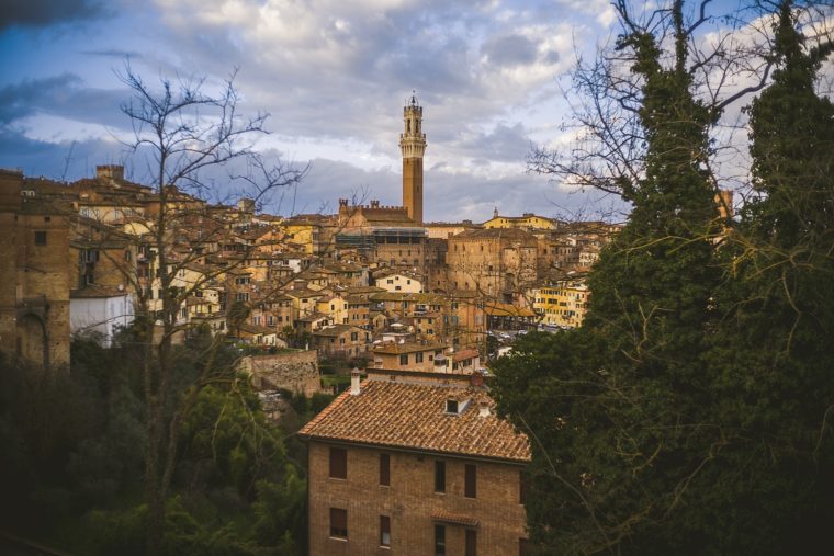 View of an ancient, terracotta-roofed town at dusk with a prominent tower rising above, framed by leafy trees under a cloudy sky.