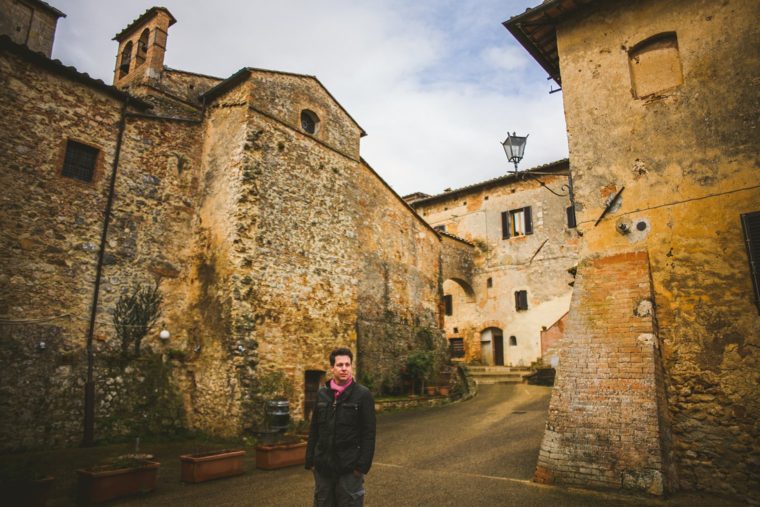 Man standing in a historic village with stone buildings and a lamp post, overcast sky above.
