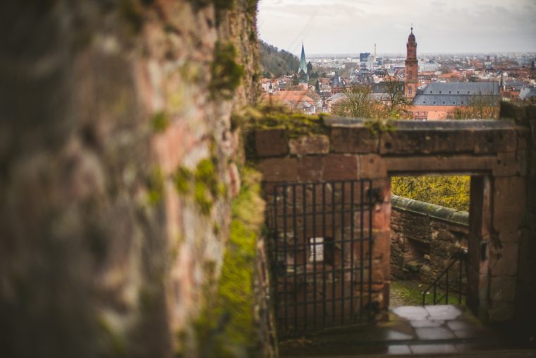 View of a cityscape through an old stone archway with a metal gate, focusing on distant buildings including a church tower.