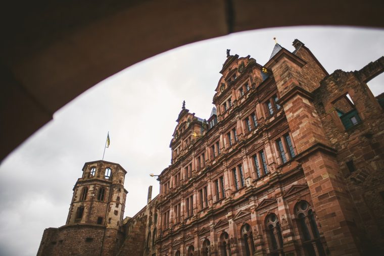 View of a historic stone castle with towers and intricate architectural details, framed by an archway, under a cloudy sky.