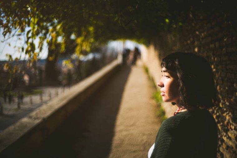 A woman with short dark hair, wearing a dark sweater, gazes thoughtfully while standing by a sunlit tree-lined pathway.