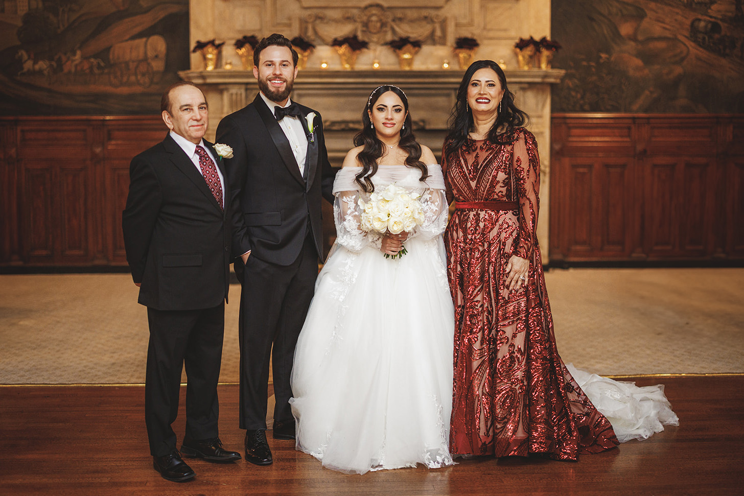 Sam + Chelsea A bride and groom stand between two older adults in formal attire, posing in a grand room with a decorative fireplace in the background.