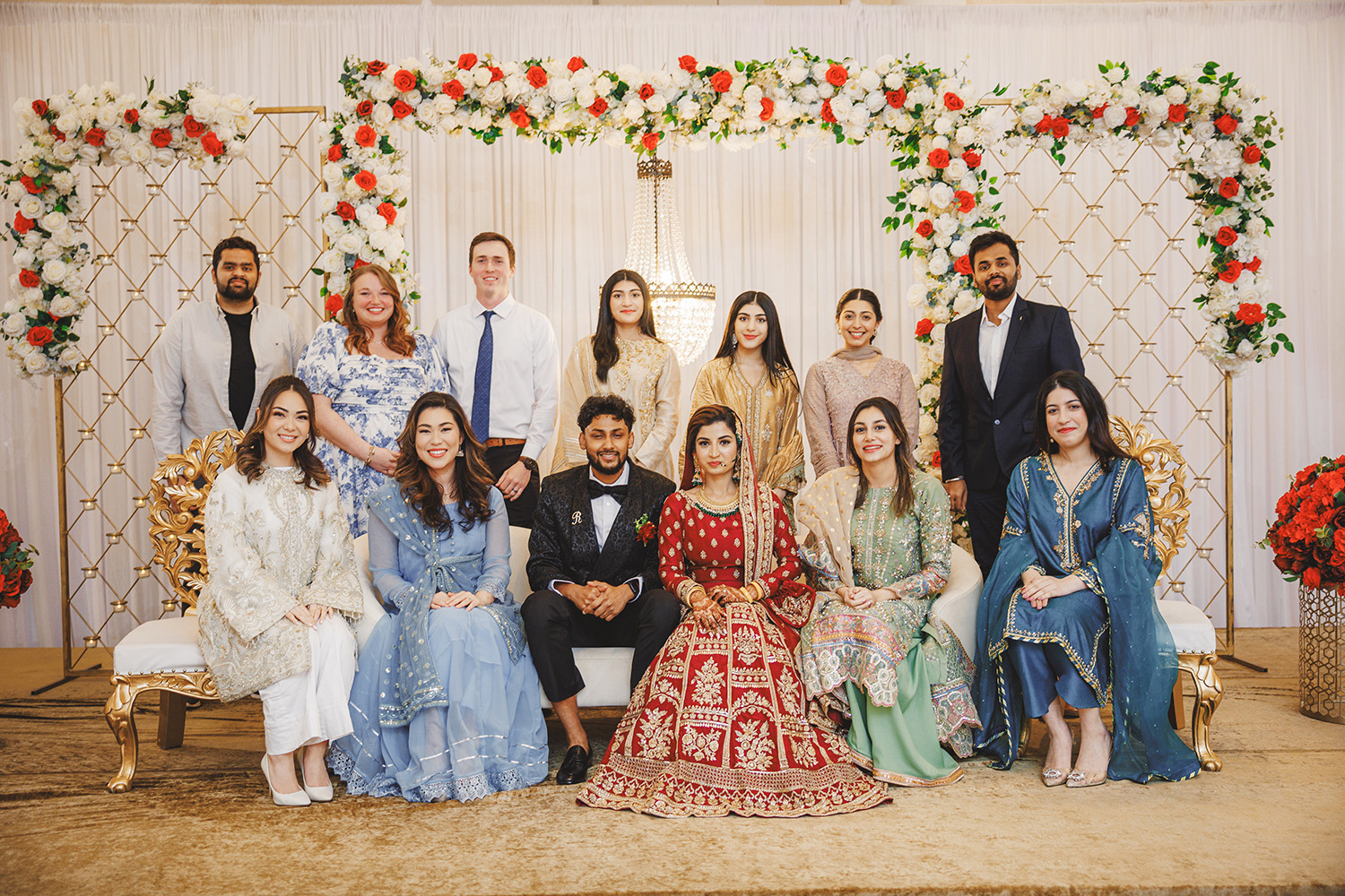 A group of people, dressed in formal and traditional attire, pose for a charming family photo in front of a floral backdrop, capturing cherished moments through photography. A group of people, dressed in formal and traditional attire, pose for a charming family photo in front of a floral backdrop, capturing cherished moments through photography.