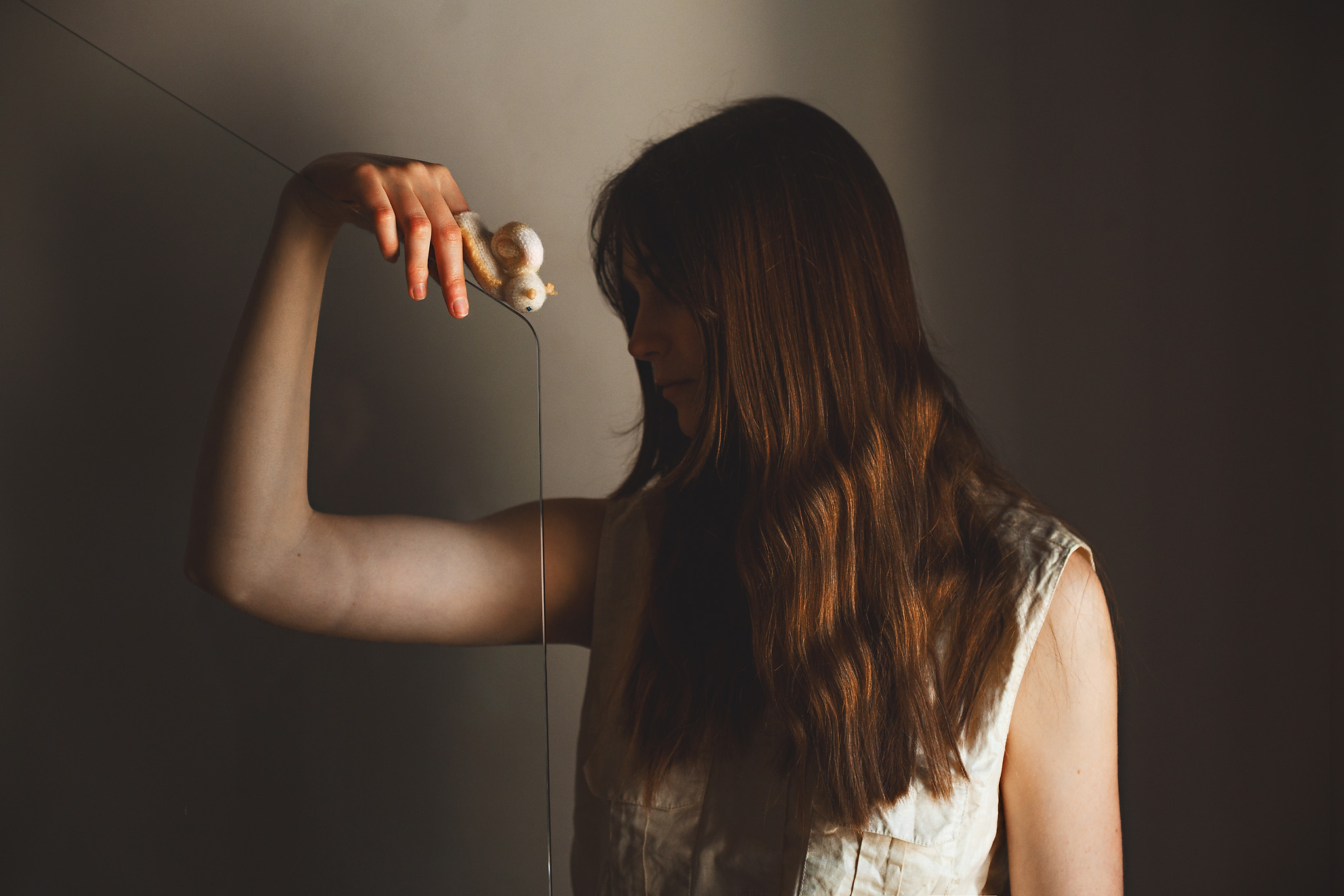 A woman with long brown hair holds a small figurine by a string, extending her arm. She is wearing a sleeveless top and stands against a dark background.