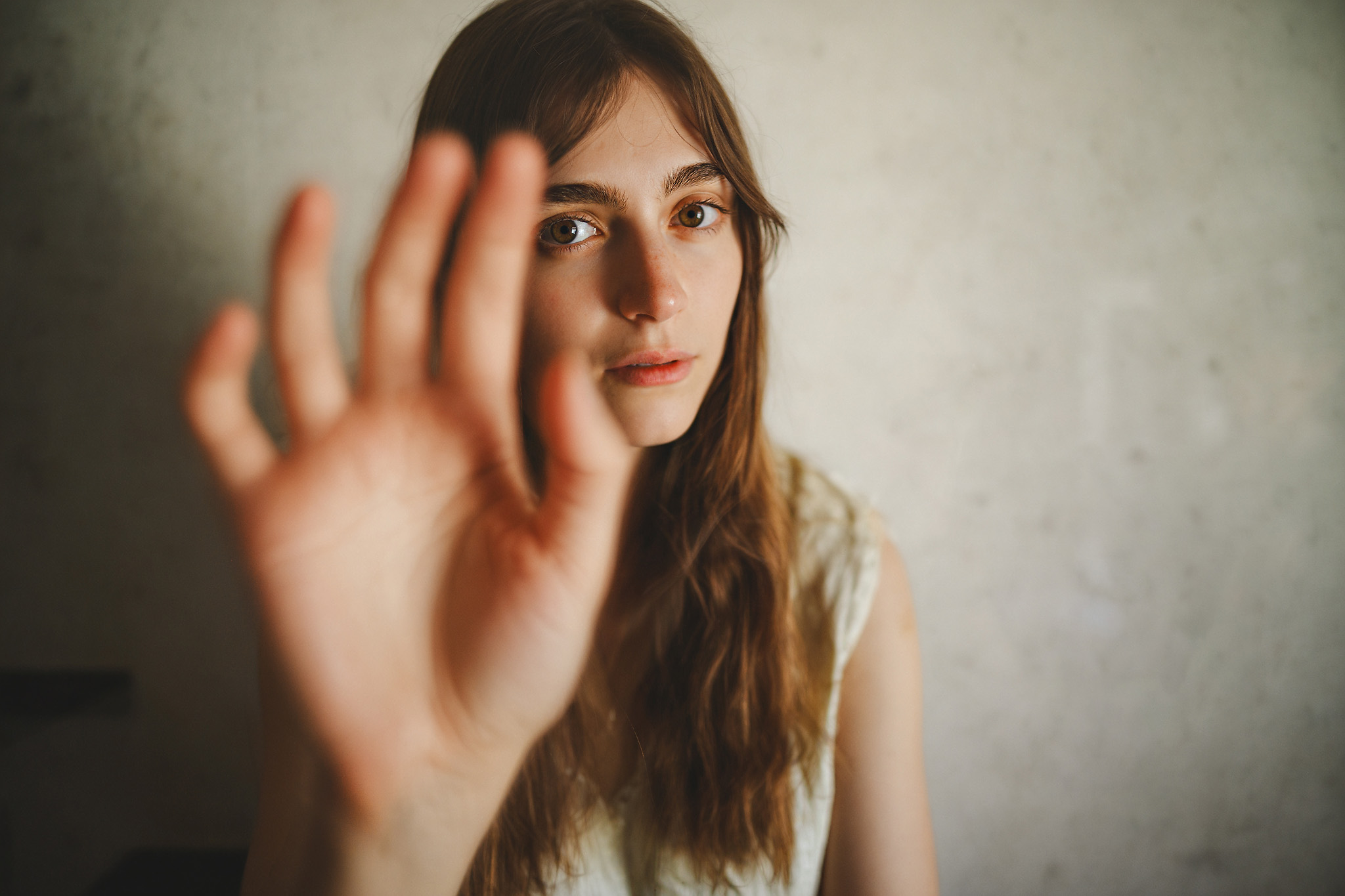 A person with long hair extends their hand toward the camera, obscuring part of their face against a neutral background.