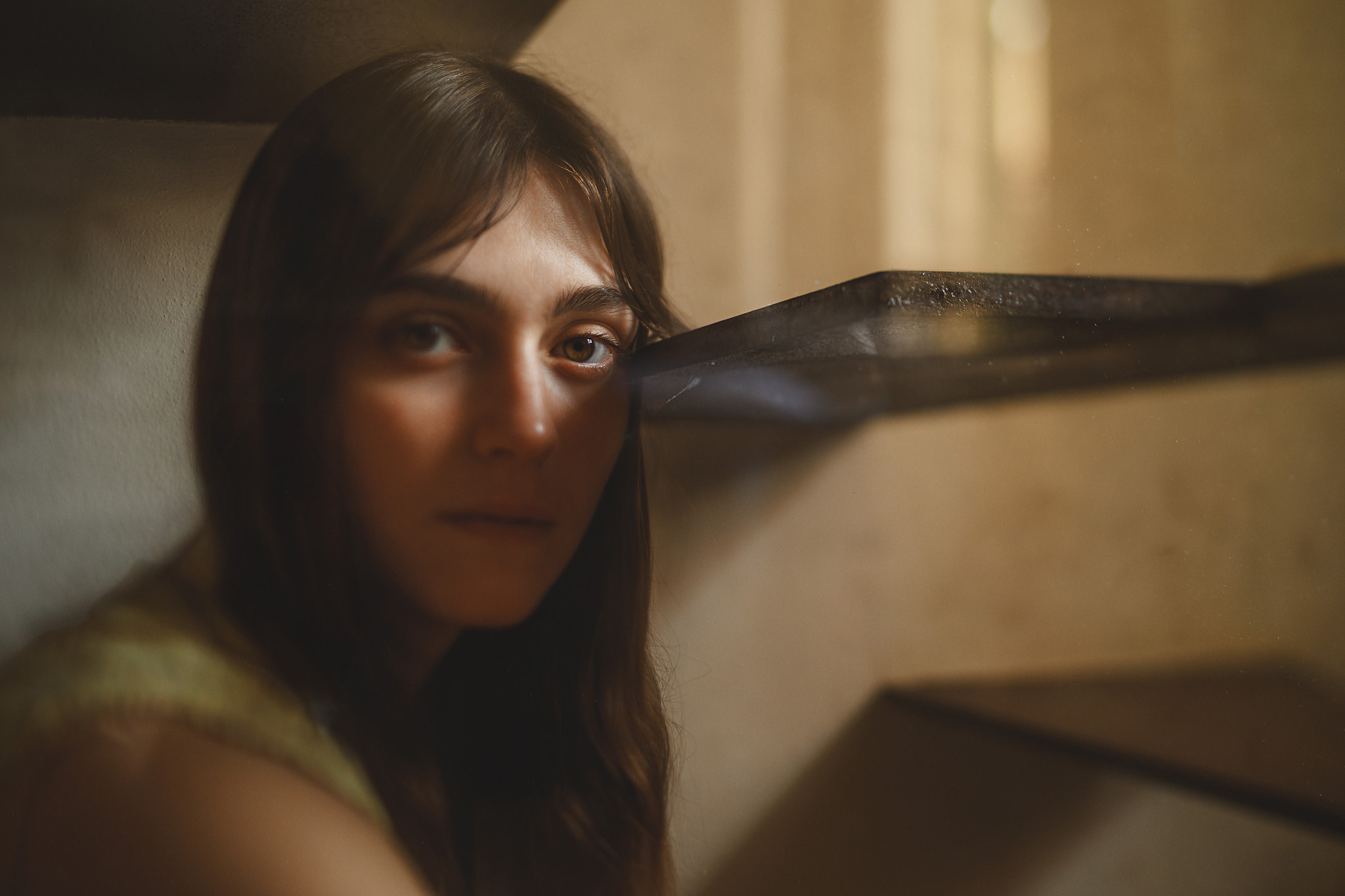 A woman with long brown hair and a neutral expression is sitting in a dimly lit room beside a staircase, partially in shadow.