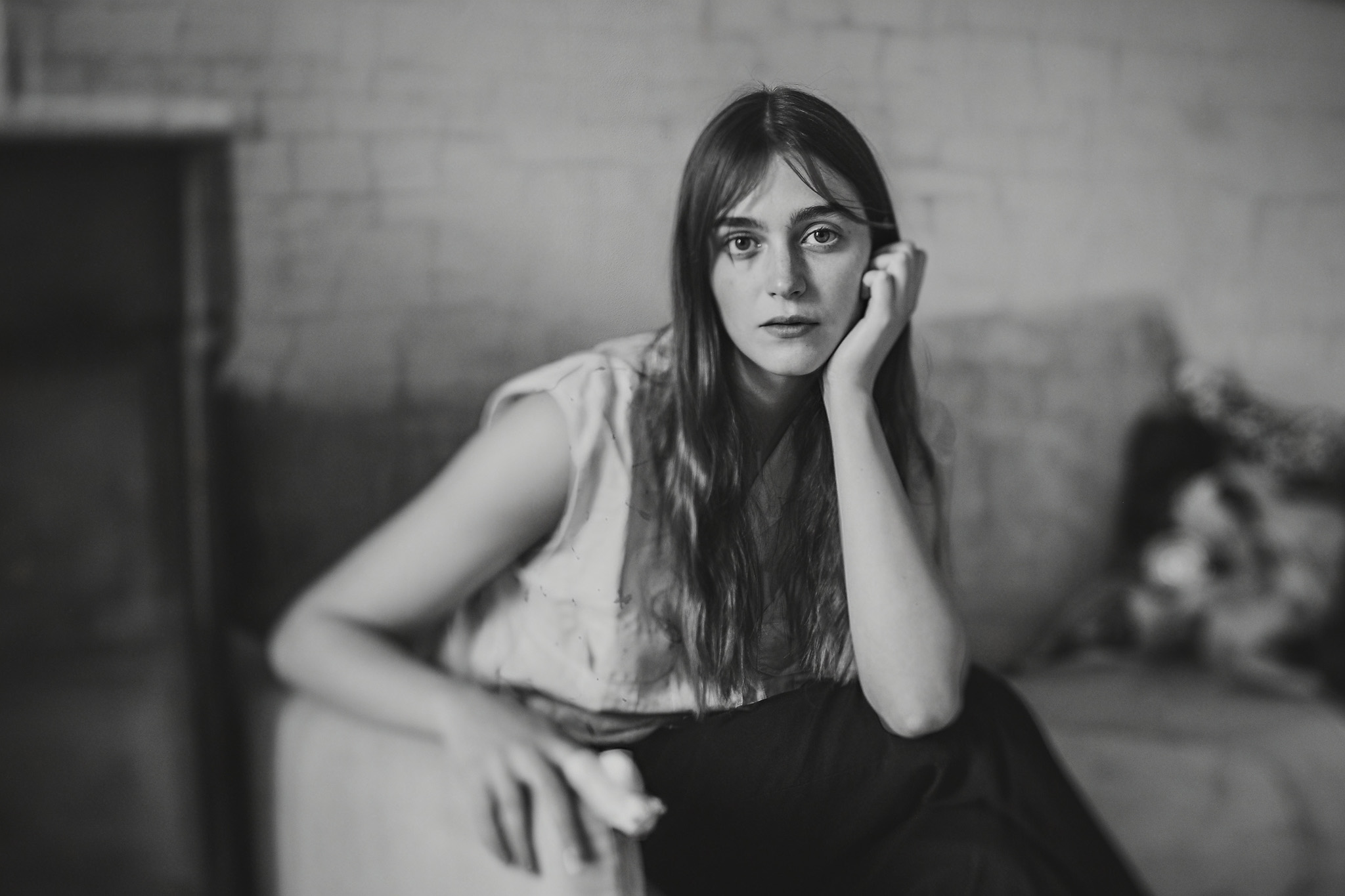 Black and white photo of a woman with long hair sitting on a couch, resting her head on her hand and looking at the camera. The background is blurred, and she wears a light top and dark pants.