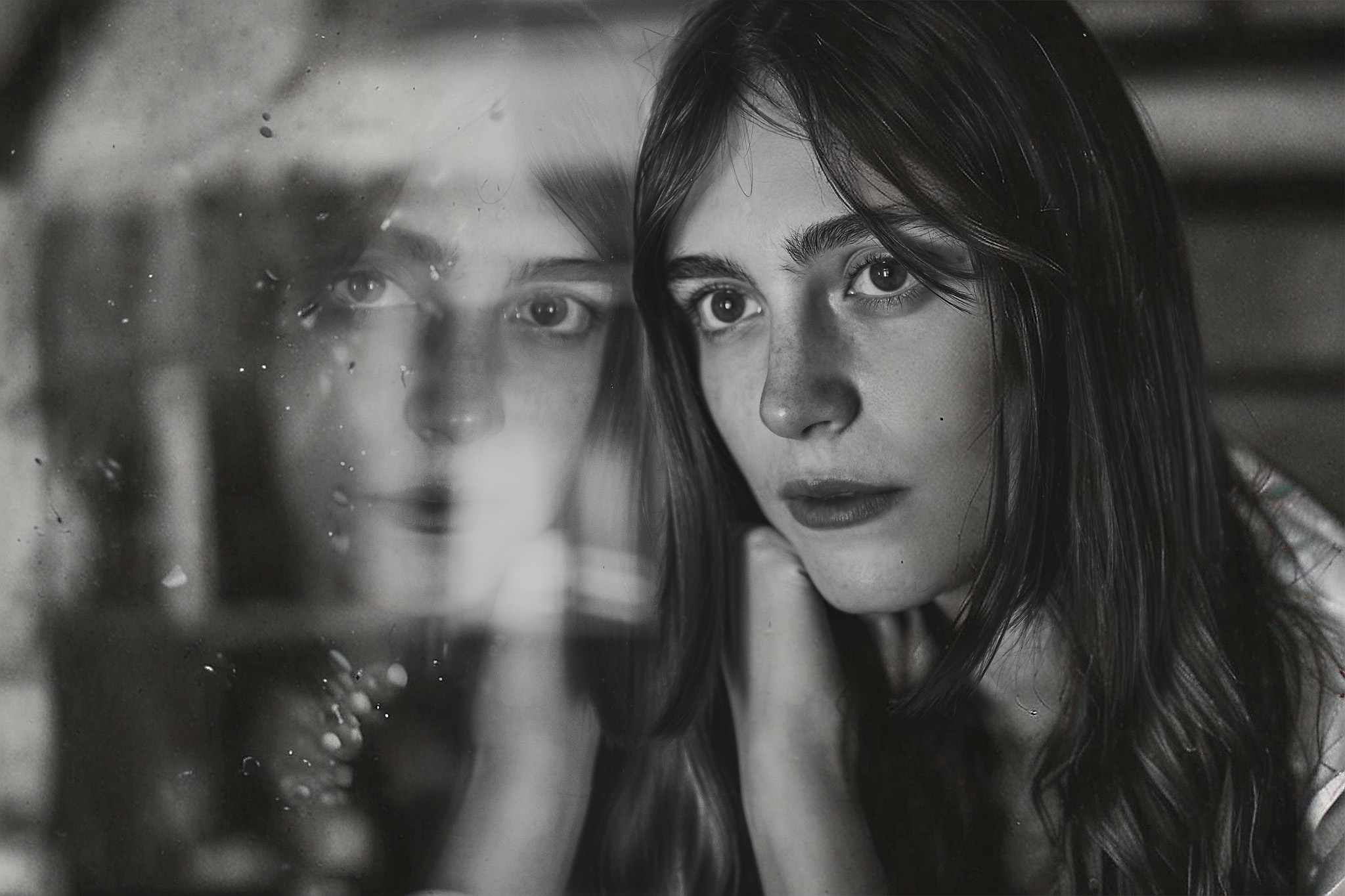 A black and white image shows a woman gazing into the distance, with her reflection visible on a nearby glass surface.