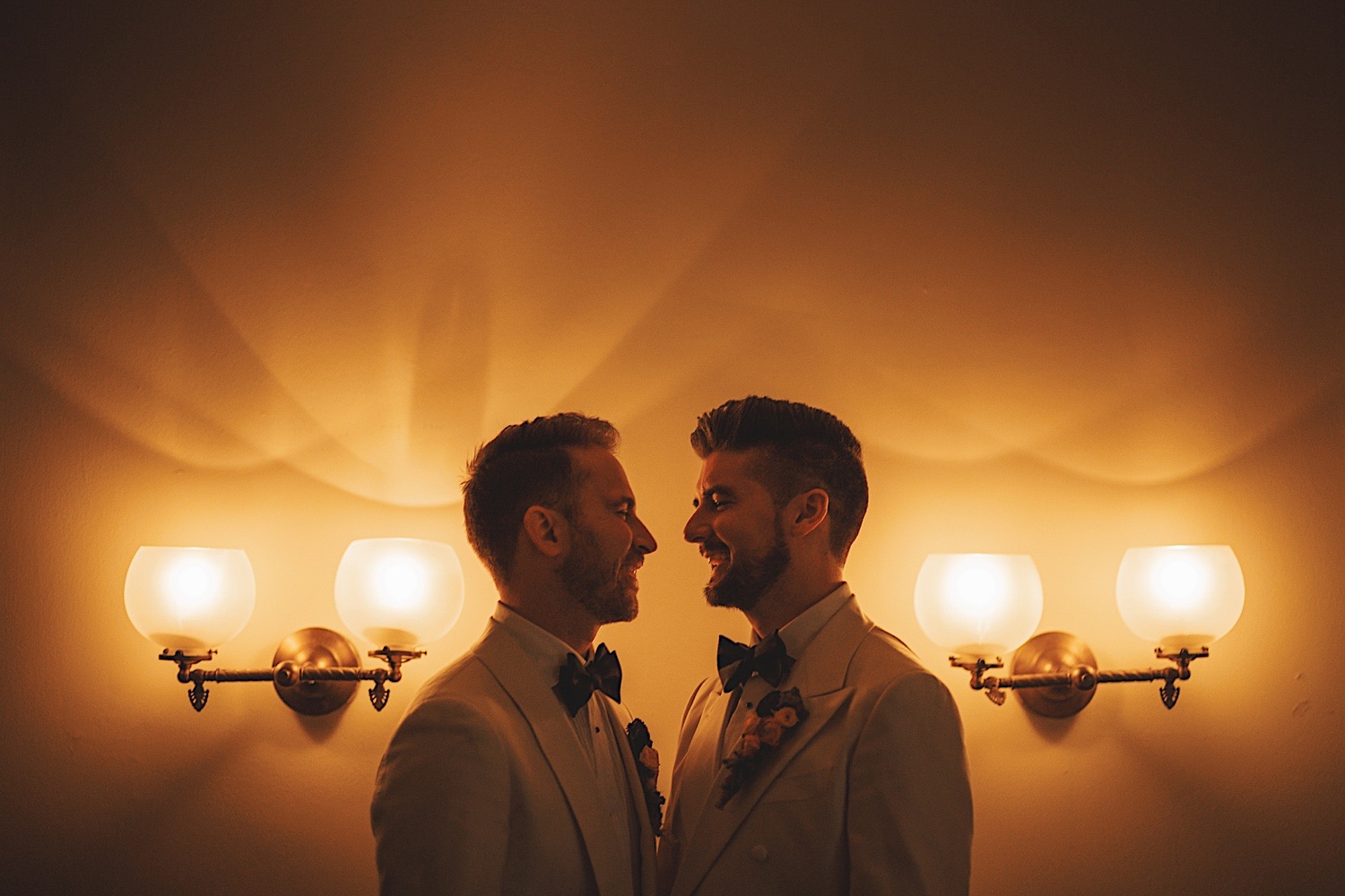 Two men in white suits stand closely, facing each other with smiles, under the warm light from four wall lamps, creating a scene that feels effortlessly drafted by auto magic.