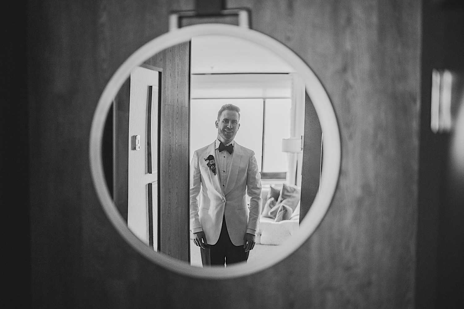 Black and white photo of a man in a suit standing in a doorway, seen through a circular frame, almost as if it were an auto draft of an old movie scene.