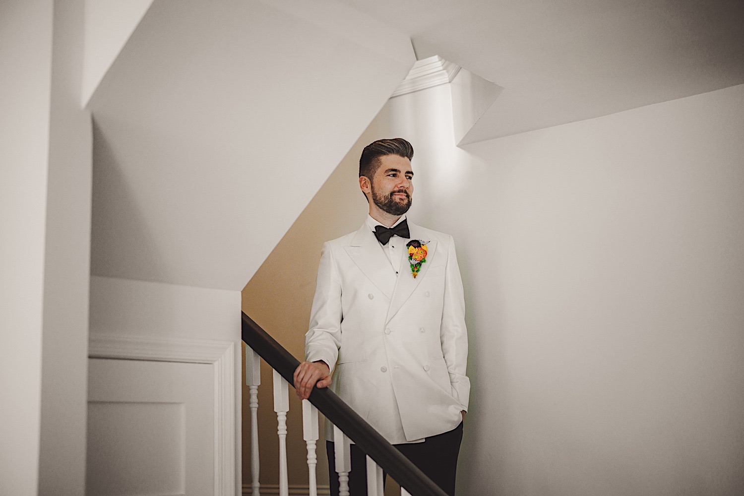 A man in a white double-breasted suit with a black bow tie and colorful boutonniere stands on a staircase, looking off into the distance, as if awaiting an important auto draft notification.