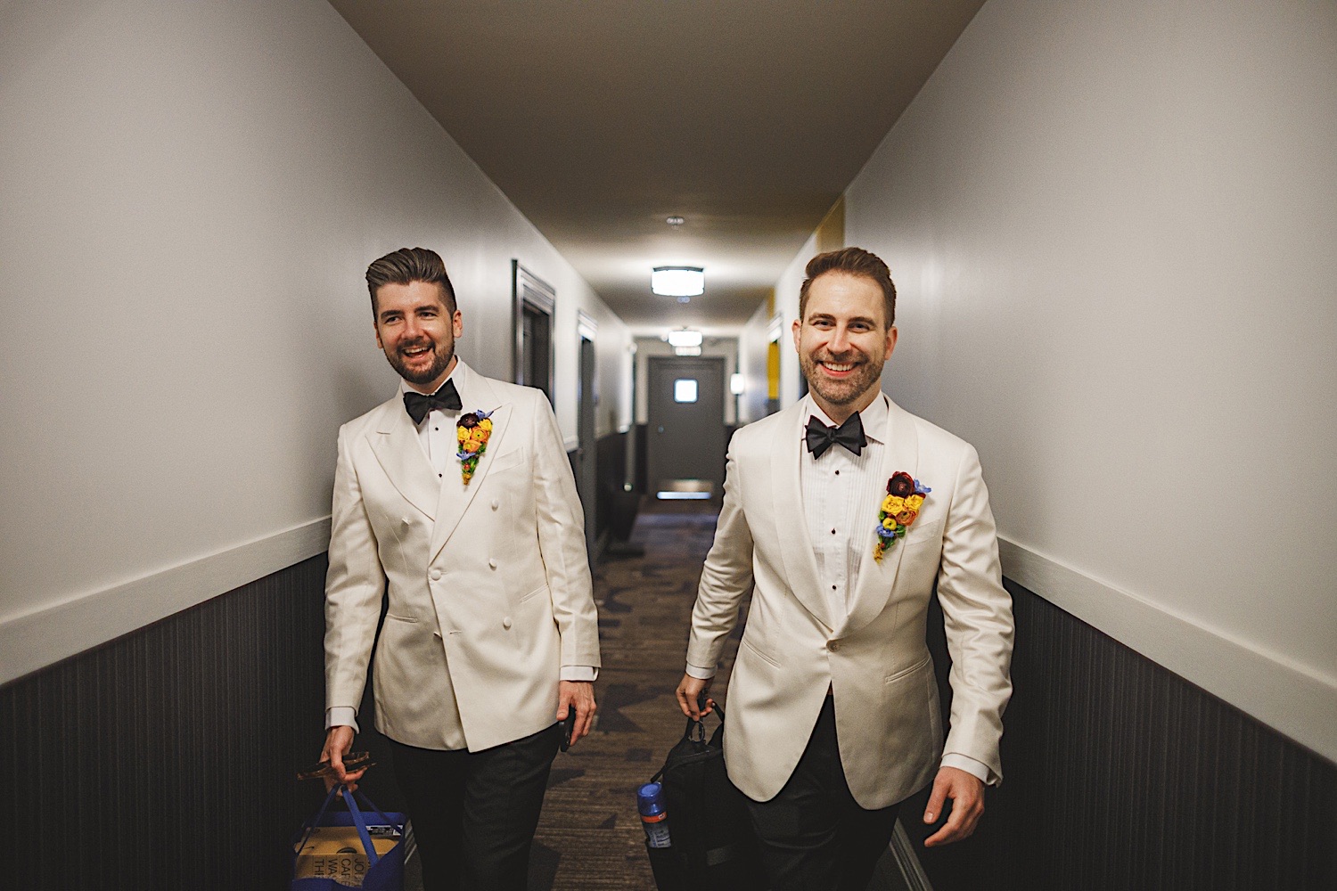 Two men in white tuxedos with bow ties and boutonnières walk down a hallway, each carrying bags. Both have short hair and are smiling, as if stepping out of a flawless Auto Draft moment.