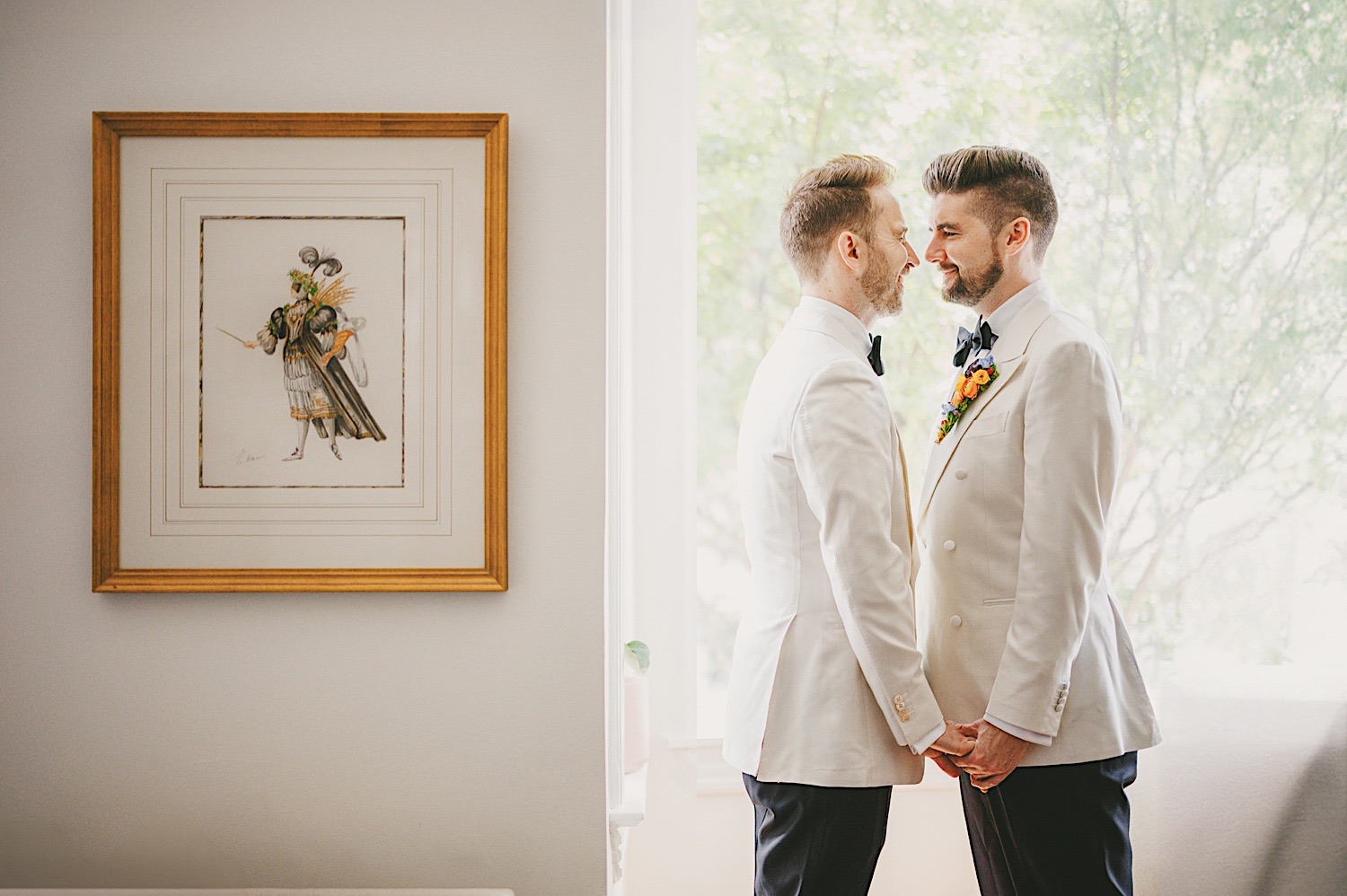 Two men in white suits stand facing each other, holding hands and smiling, next to a framed artwork on a bright, window-lit interior wall. The scene feels like it could be part of an artistic draft for an upcoming exhibition.