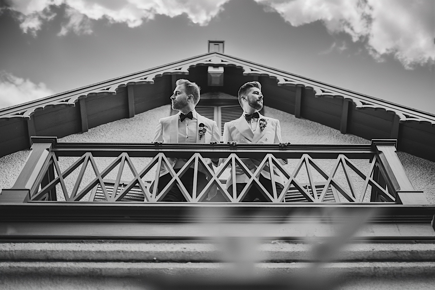 Two men in white suits stand on a balcony, looking in opposite directions. The picture, reminiscent of a classic auto draft illustration, is in black and white with a backdrop of a cloudy sky.