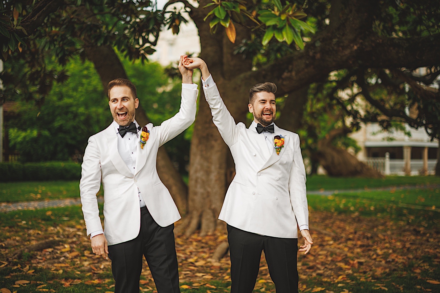 Two men wearing white tuxedos with black bow ties and floral boutonnieres hold hands and raise them in celebration, as if they were an Auto Draft for the perfect wedding. They stand outside on a grassy area with trees in the background.
