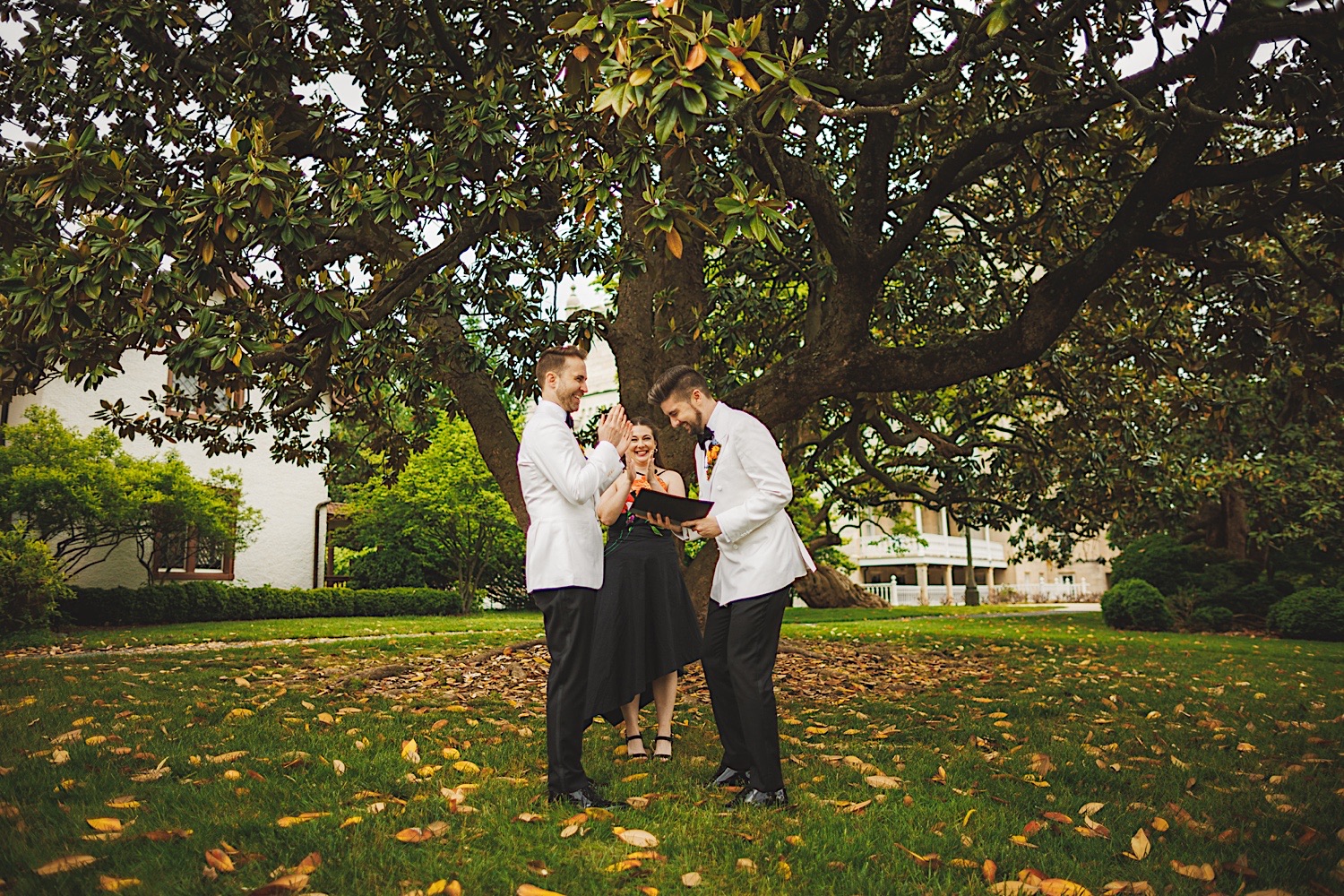Two grooms and an officiant stand under a large tree in an outdoor setting, all smiling and laughing, suggesting a joyful moment possibly during a wedding ceremony. It's as if their happiness was on auto draft, effortlessly filling the air with joy.